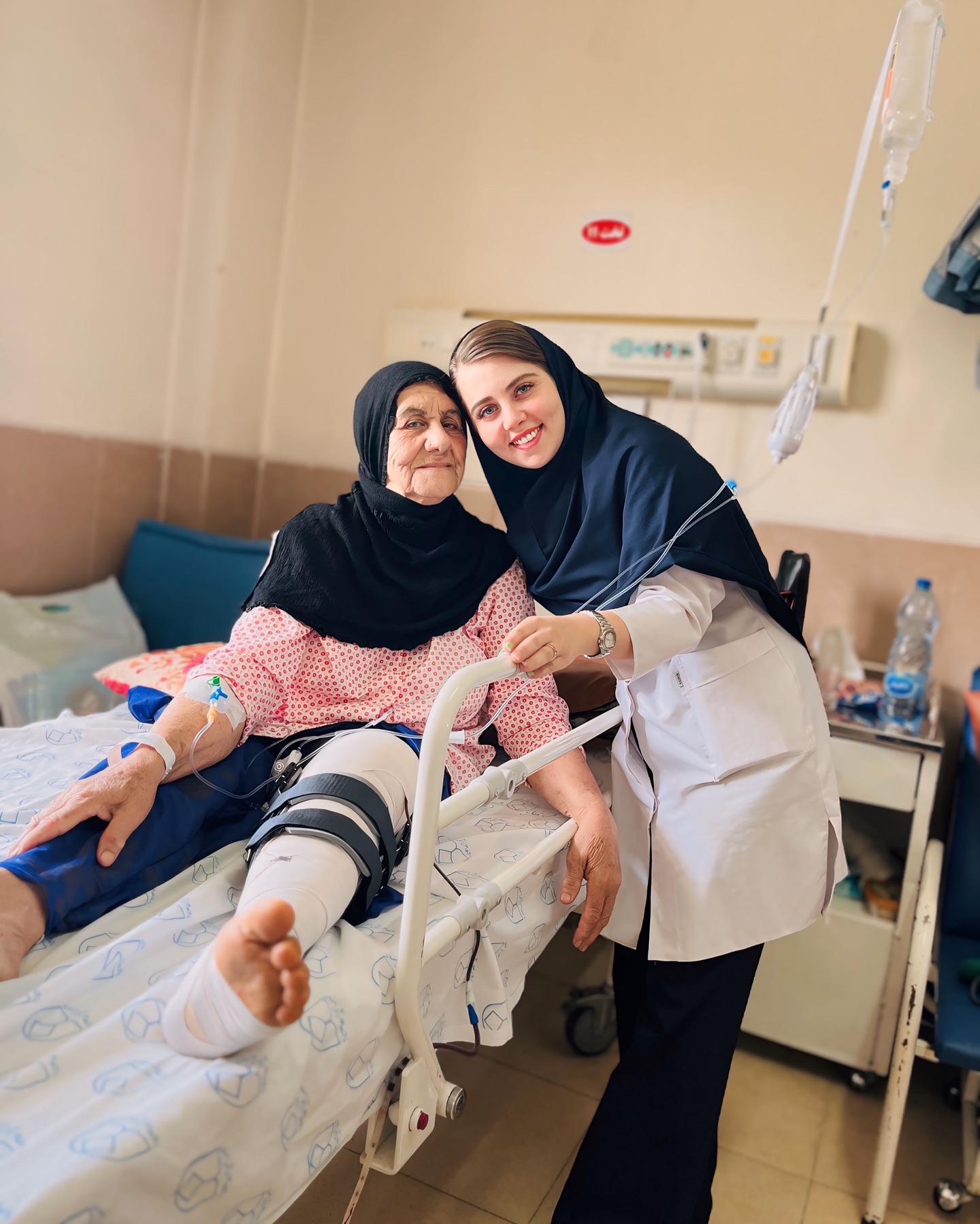  یک خانم سالمند روی تخت بیمارستان نشسته و پایش بانداژ و آتل شده است. او سرم به دست دارد و به نظر می‌رسد تحت مراقبت پزشکی قرار دارد. کنار او یک پرستار یا کارمند درمانی با روپوش سفید و مقنعه تیره ایستاده و لبخند می‌زند. هر دو با هم ارتباط خوبی دارند و فضای عکس صمیمانه و دلگرم‌کننده است. 